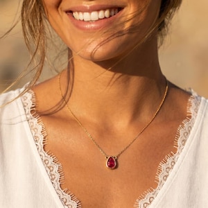 May include: A gold necklace with a teardrop-shaped, red gemstone pendant. The pendant is set in a gold bezel. The necklace is worn over a white top with lace trim. The image is a close-up of the necklace and the person wearing it.