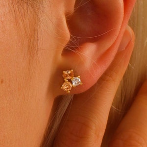 Puede incluir: Pendientes de oro con tres pequeñas piedras, dos son de color melocotón y una es transparente.