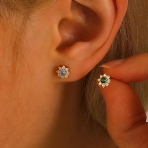 May include: A pair of gold flower-shaped earrings with a blue and green gemstone in the centre. The earrings are surrounded by smaller clear gemstones.