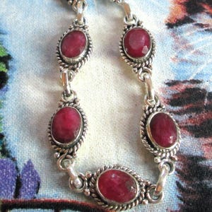 Puede incluir: Una pulsera de plata con siete piedras preciosas de rubí rojo intenso, de forma ovalada y facetadas. Cada piedra está engastada en un marco de plata trenzada y unida por eslabones de plata. La pulsera incluye un cierre de barra en el extremo.