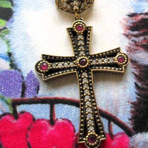 Puede incluir: Un collar con colgante de cruz de estilo vintage. La cruz es negra con ribetes dorados, adornada con piedras preciosas claras y rojas. El collar tiene una delicada cadena dorada y una argolla decorativa.