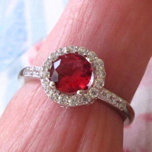 May include: A silver ring featuring a large, round, deep red gemstone surrounded by a halo of smaller, clear gemstones. The ring's band is also adorned with small, clear gemstones, creating a sparkling effect. The ring is a piece of jewelry.