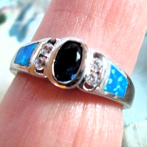 May include: A silver ring featuring a large, oval, dark blue gemstone at the center, flanked by smaller clear stones and blue opal-like inlays. The ring is set against a skin-toned background, highlighting its intricate design and vibrant colors.