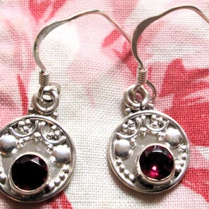May include: A pair of silver-toned earrings with a circular design. Each earring features a deep red, round gemstone at the center, surrounded by a decorative border with small silver accents. The earrings have a hook closure.