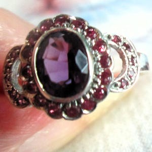 May include: A silver ring featuring a large, oval-cut purple center stone surrounded by a halo of small, round, deep red stones. The band includes additional small red stones set into the metalwork on both sides of the central cluster setting.