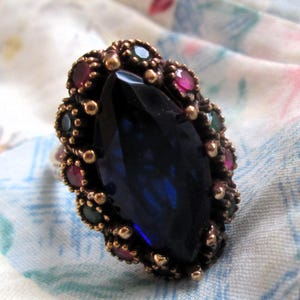 May include: A ring featuring a large, central marquise-cut dark blue stone surrounded by a cluster of smaller, round-cut red and green stones set in a textured, dark metallic frame that creates a dense, ornate border around the main gemstone.