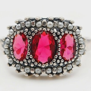 May include: A silver ring featuring three oval, vibrant pink gemstones. The gemstones are surrounded by a border of small, silver-colored beads. The ring has a vintage or antique aesthetic.