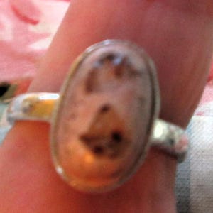 May include: A silver ring with an oval, light-colored stone featuring brown and black inclusions. The ring is set on a finger, showcasing the design and details of the jewelry.