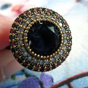 May include: A large, ornate ring with a dark blue center stone. The ring is surrounded by multiple rows of smaller stones in white, red, green, and blue, set in a gold-colored metal. The ring's design is intricate and eye-catching.