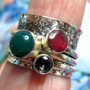 May include: A wide silver spinner ring featuring an embossed floral pattern on the band. The ring is adorned with three bezel-set gemstones: a large round smooth green stone, a smaller round faceted red stone, and a small round faceted dark stone.