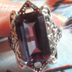 May include: A silver-toned ring featuring a large, rectangular, deep purple faceted gemstone as the centerpiece. The metal setting displays intricate scrollwork and filigree patterns, accented by small, vibrant red stones at the top and bottom.