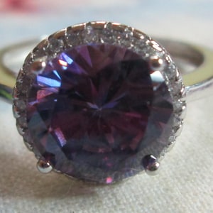 May include: A silver-toned ring featuring a large, round, faceted purple center stone. The central stone is surrounded by a halo of small, clear, sparkling stones set into the silver-toned metal band, which has a smooth, polished finish.