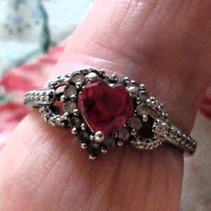 Puede incluir: Anillo plateado con una piedra roja en forma de corazón. El anillo presenta pequeñas piedras claras y detalles intrincados alrededor del corazón y la banda, creando una estética vintage o romántica, ideal para ocasiones especiales.