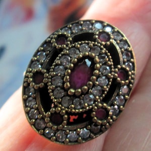 May include: An oval-shaped ring featuring a central marquise-cut deep red stone surrounded by a halo of small clear stones. The setting is made of a dark, antiqued metal with additional red and clear stones arranged in a decorative pattern.