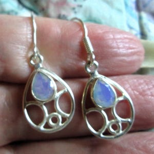 May include: A pair of silver-toned drop earrings. Each earring features a teardrop-shaped, iridescent, pale blue gemstone set within an openwork, decorative frame. The earrings have a hook closure.