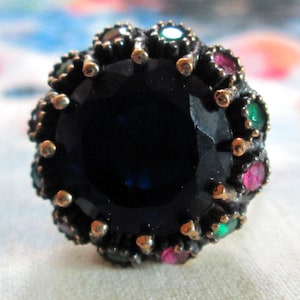 May include: A dark metal ring featuring a large, round, faceted deep blue central stone. The center stone is surrounded by a halo of smaller, alternating round faceted stones in vibrant shades of emerald green and ruby red, set in a dark metal frame.