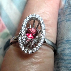 May include: A silver ring with an oval-shaped setting. The setting is encrusted with small, clear stones and surrounds a vibrant red gemstone. The ring is on a finger, showcasing its design and sparkle.