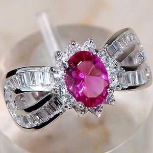 May include: A silver-toned ring featuring a central oval-cut vibrant pink gemstone surrounded by a halo of small clear stones. The band is shaped like a bow and is embellished with rows of rectangular-cut clear stones on both sides.