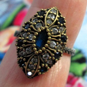 Puede incluir: Un anillo ornamentado con una piedra azul ovalada central, rodeada de gemas claras y azul oscuro. El anillo tiene un diseño de filigrana dorada y una banda plateada. Una pieza de joyería.