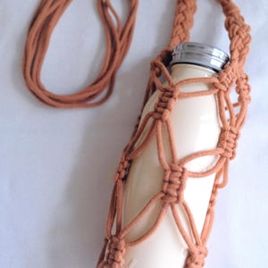 Pu&ograve; includere: Un porta bottiglia in crochet marrone con una bottiglia d'acqua bianca all'interno. Il porta bottiglia ha una lunga tracolla per portarlo sulla spalla.