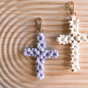 Pu&ograve; includere: Due portachiavi a forma di croce realizzati a uncinetto, uno bianco e uno viola. I portachiavi sono appesi a una superficie di legno.