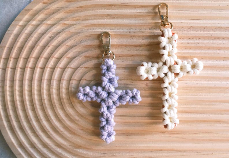 Pu&ograve; includere: Due portachiavi a forma di croce realizzati a uncinetto, uno bianco e uno viola. I portachiavi sono appesi a una superficie di legno.