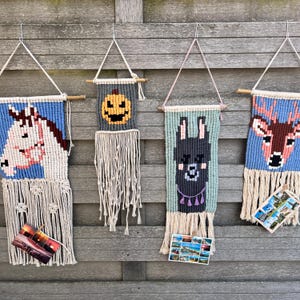 May include: Four colourful wall hangings with different pixelated animal designs. The designs include a unicorn, a pumpkin, a llama, and a deer. Each hanging has a wooden dowel at the top and white yarn fringe at the bottom.