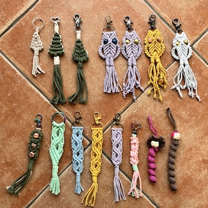 May include: A collection of 18 handmade macrame keychains featuring various designs, including owls, trees, and geometric patterns. The keychains are made with colorful yarn and have silver metal clasps.