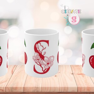 May include: White ceramic mug featuring a pair of red cherries with green leaves. The mug on the left has the name "Susana" in red script, and the mug on the right has the word "cherry". The middle mug has the letter "S" with floral accents.