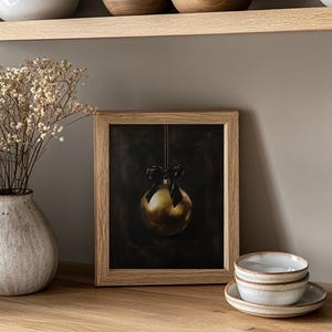 May include: A framed print of a gold Christmas ornament with a black ribbon hanging from a string against a dark background.