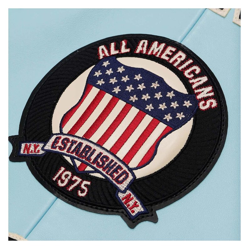 May include: A circular patch with the text "ALL AMERICANS" in red embroidery. The patch features a shield with the American flag design, a banner reading "ESTABLISHED 1975 N.Y.", and a black border. The background is light blue.