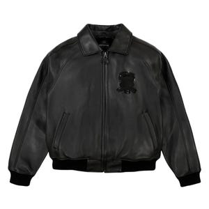 Könnte beinhalten: Schwarze Leder-Bomberjacke mit Kragen, Reißverschluss und zwei Vordertaschen. Die Jacke hat ein schwarzes Emblem auf der Brust und schwarze gerippte Bündchen und Saum. Der Markenname "AVIREX" ist auf dem Kragen sichtbar.