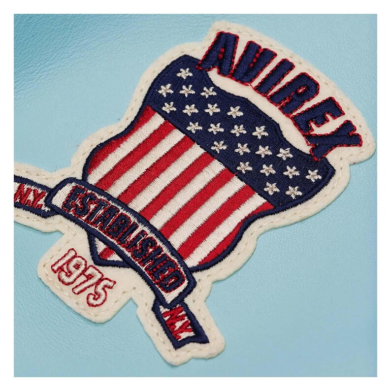 May include: Embroidered patch featuring a shield design with the American flag and the text "AVIREX ESTABLISHED 1975 NY". The patch has a white border and is set against a light blue background.