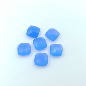 May include: Six light blue, square-cut gemstones with faceted surfaces are arranged on a white background. The gemstones have rounded corners and a translucent appearance, reflecting light. These could be used for jewelry making.