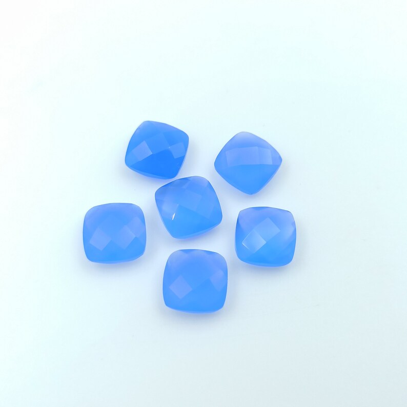 May include: Six light blue, square-cut gemstones with faceted surfaces are arranged on a white background. The gemstones have rounded corners and a translucent appearance, reflecting light. These could be used for jewelry making.