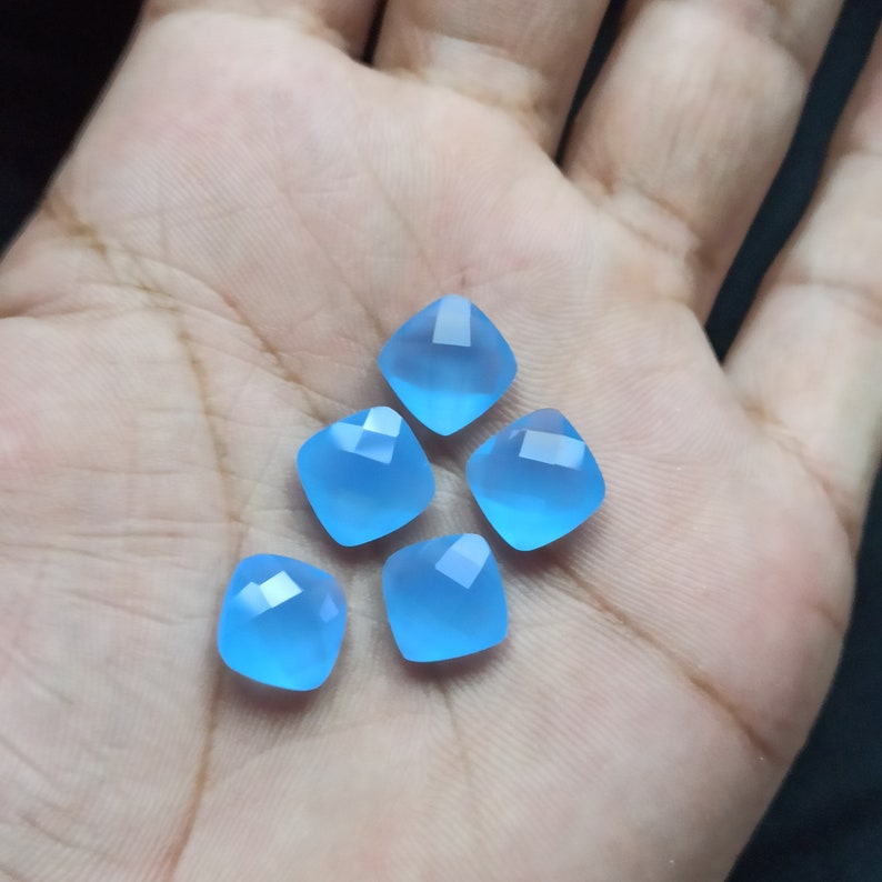 May include: Five light blue, square-cut gemstones with faceted edges. The gems are a soft, translucent blue and are arranged in a hand. The gemstones have a cushion cut and are likely for jewelry making.