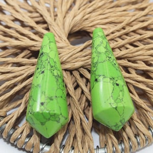 Pode incluir: Dois pingentes de pedras preciosas verdes brilhantes, facetadas, com veios pretos. As pedras em forma de gota são exibidas em um fundo circular tecido. Os pingentes são provavelmente para fazer joias ou artesanato.