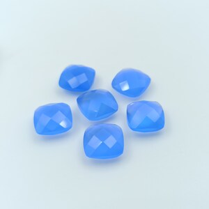 May include: Six light blue, square-cut gemstones are arranged on a white surface. Each gem has multiple facets that reflect light, creating a sparkling effect. The gems are a soft, pastel blue color.