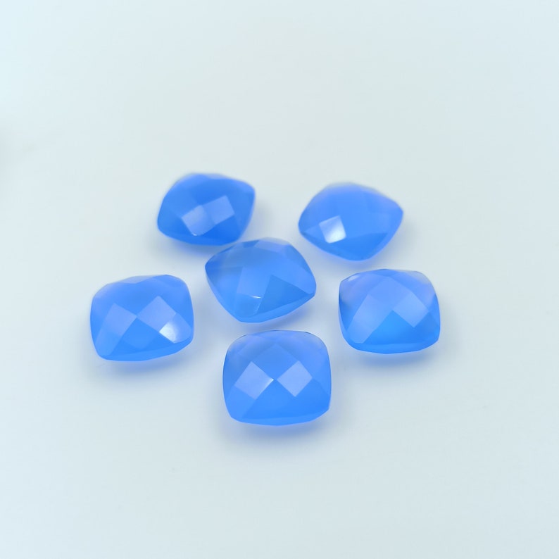 May include: Six light blue, square-cut gemstones are arranged on a white surface. Each gem has multiple facets that reflect light, creating a sparkling effect. The gems are a soft, pastel blue color.
