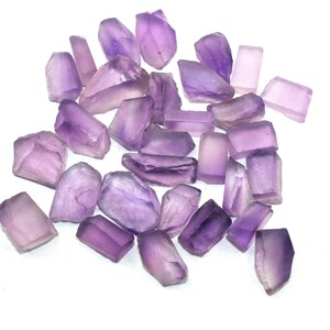 May include: A collection of raw amethyst gemstones in various shapes and sizes. The stones display shades of purple and lavender, with a translucent quality. The gemstones are scattered against a white background, showcasing their natural, unpolished state.