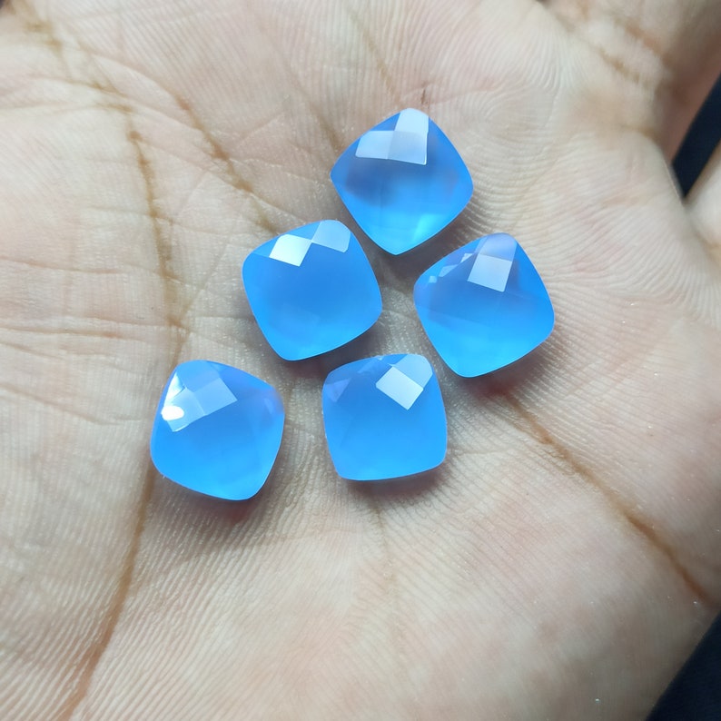 May include: Five square-cut, light blue gemstones with faceted tops. The gems are arranged on a light-colored surface, showcasing their clarity and color. The gems are a vibrant, translucent blue.
