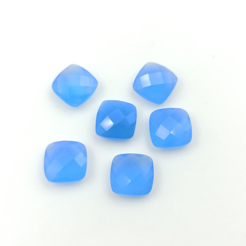 May include: Six light blue, square-shaped gemstones with faceted surfaces are scattered on a white background. The gemstones have a soft, translucent appearance, and their cut creates multiple reflective surfaces.