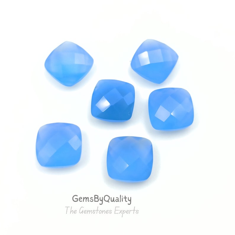 May include: Six light blue, square-cut gemstones with faceted surfaces are arranged on a white background. The gemstones have rounded corners and a translucent appearance. The text "GemsByQuality" and "The Gemstones Experts" is visible.