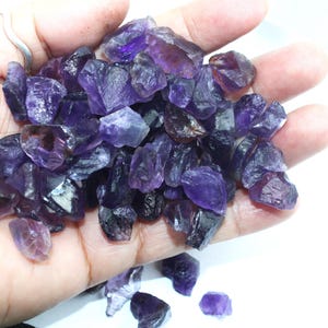 May include: A handful of raw amethyst crystals in varying shades of purple and violet. The rough-cut gemstones have a natural, unpolished appearance, with visible facets and textures. The crystals are displayed against a bright white background.