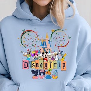 Disney Trip 2026 hoodie with Mickey Mouse, Minnie Mouse and Disney friends in front of a colorful castle with fireworks inside Minnie ears silhouette on blue pullover hoodie.