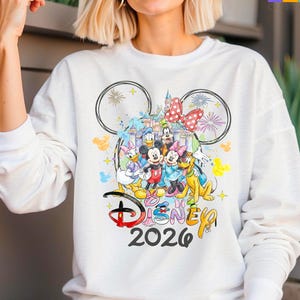 May include: White long-sleeved sweatshirt with a vibrant Disney-themed graphic. The design showcases Mickey and Minnie Mouse, Donald and Daisy Duck, and Goofy, alongside a castle, fireworks, and the text "Disney 2026".
