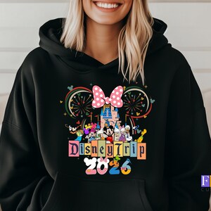 Puede incluir: Sudadera con capucha negra con un gr&aacute;fico colorido de personajes de Disney, un castillo, fuegos artificiales y un lazo rosa. El texto "Disney Trip 2025" se muestra en una fuente vibrante y multicolor.