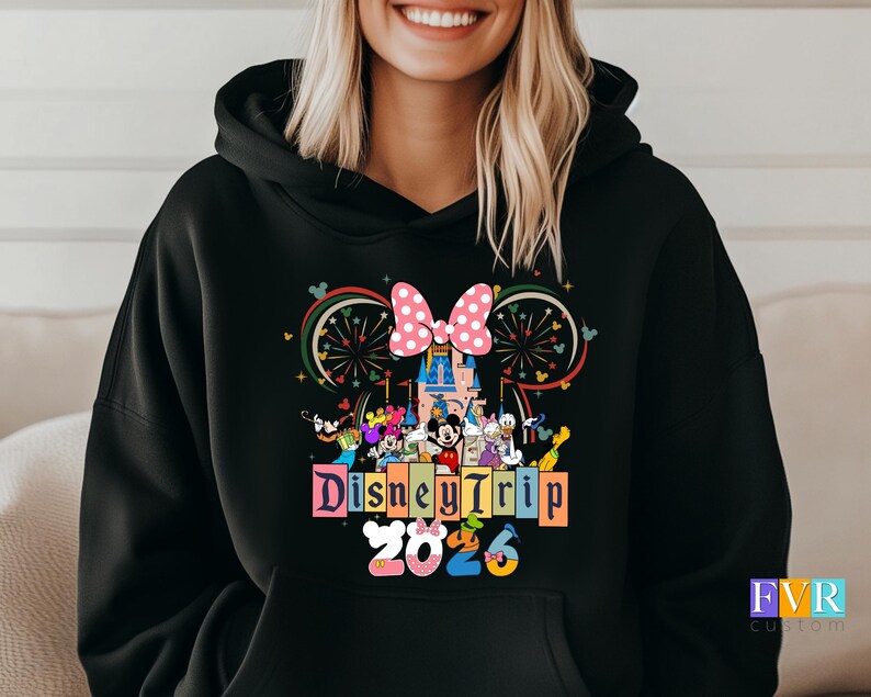 Puede incluir: Sudadera con capucha negra con un gr&aacute;fico colorido de personajes de Disney, un castillo, fuegos artificiales y un lazo rosa. El texto "Disney Trip 2025" se muestra en una fuente vibrante y multicolor.