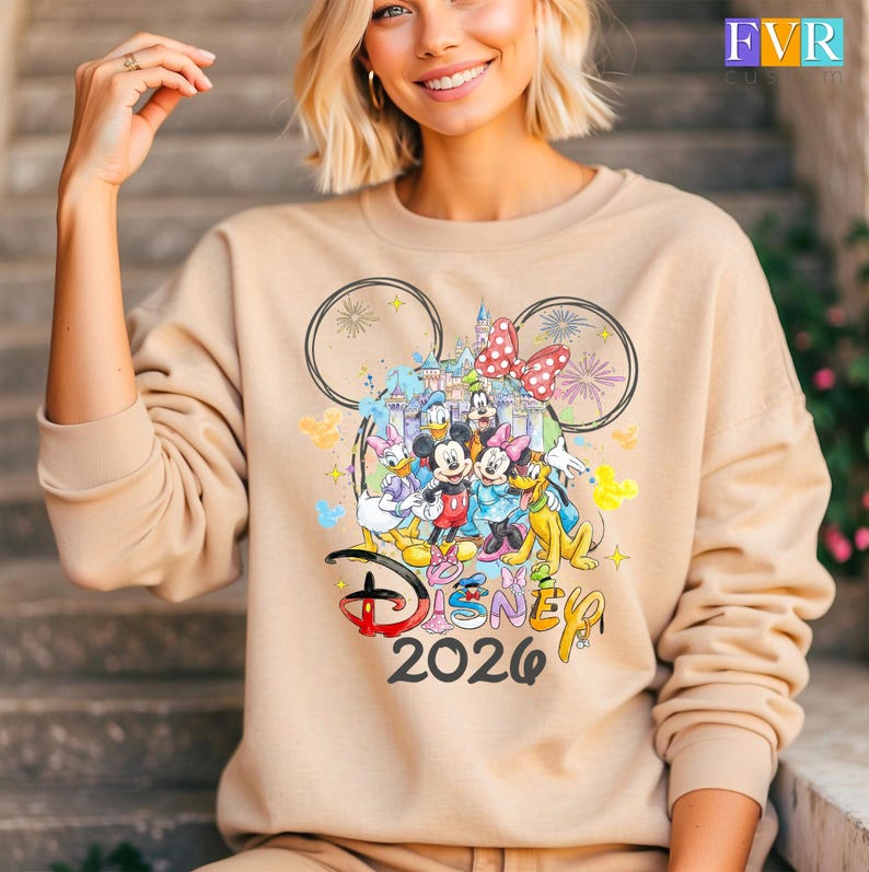 May include: Beige sweatshirt with a Disney design. The design includes Mickey Mouse, Minnie Mouse, Donald Duck, Daisy Duck, Goofy, and Pluto, a castle, fireworks, and the text "Disney 2026".