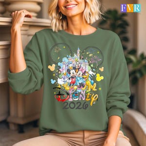 May include: An olive green sweatshirt with a vibrant Disney design. The graphic showcases Mickey Mouse, Minnie Mouse, Donald Duck, and other characters, a castle, fireworks, and the text "Disney 2026".